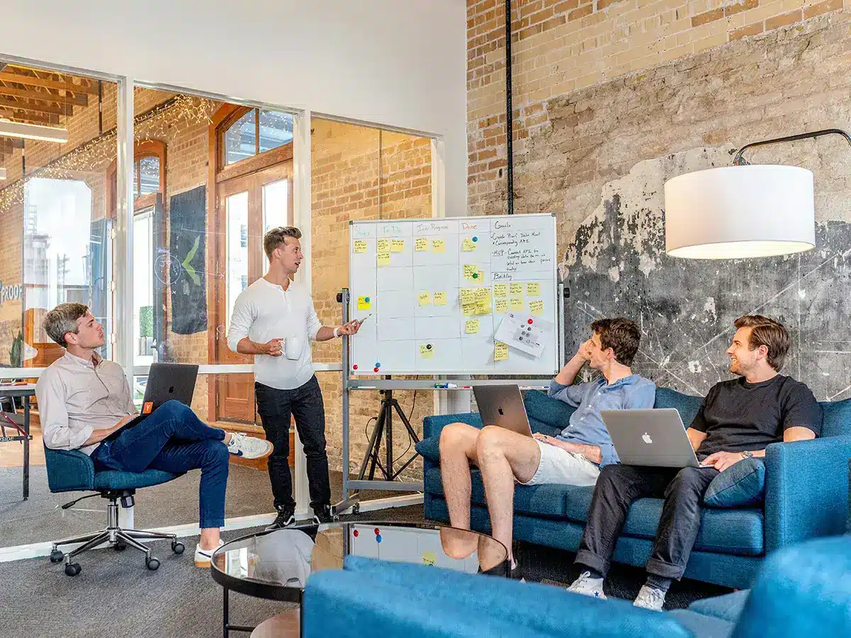 Four people in a casual office setting. One person stands, pointing to a whiteboard with sticky notes, while the other three sit on couches with laptops. The room has exposed brick walls and large windows in the background.