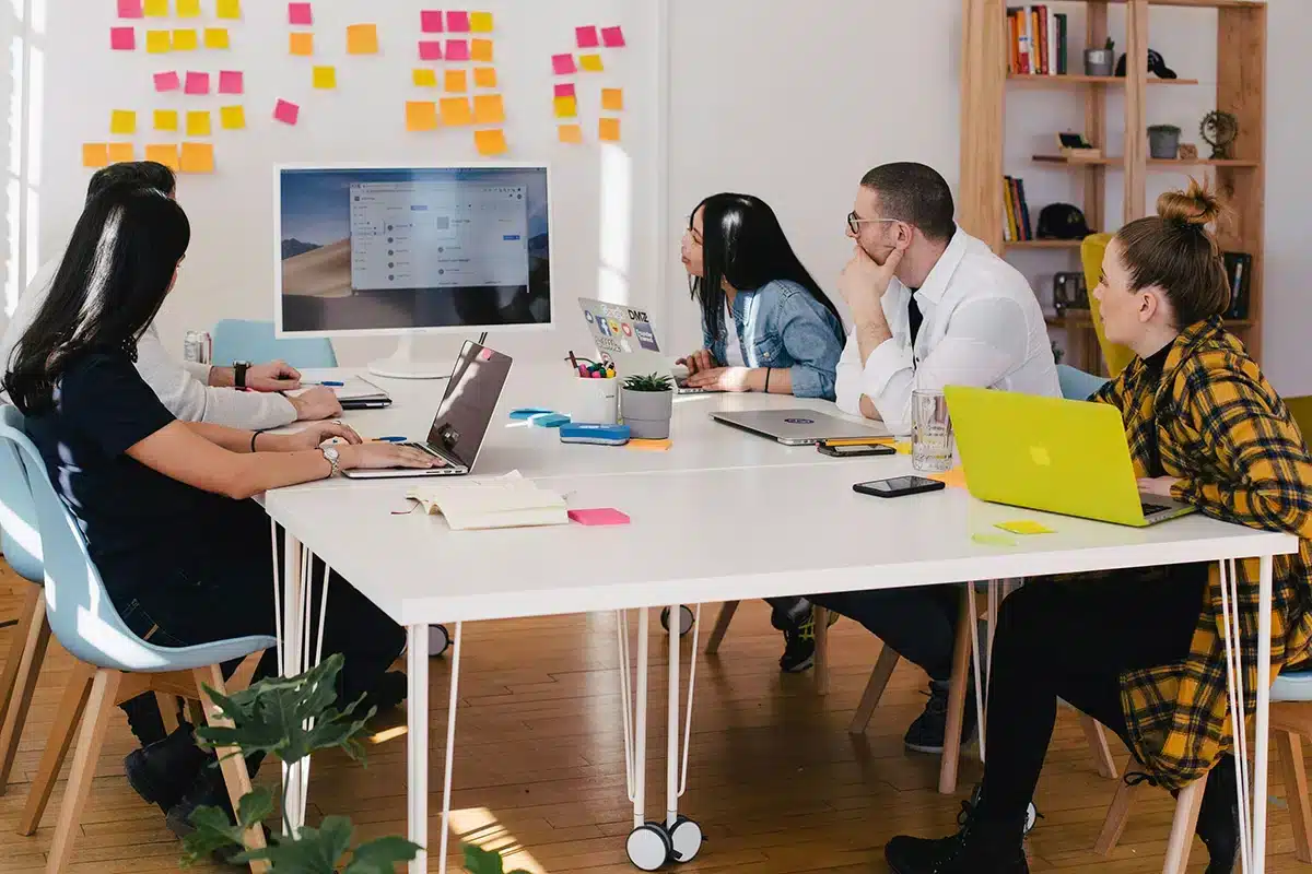 A group of five people sit at a table in a modern office, focused on a presentation displayed on a wide monitor. Laptops, colorful sticky notes, and office supplies are on the table. Shelves and a wall adorned with sticky notes are in the background.
