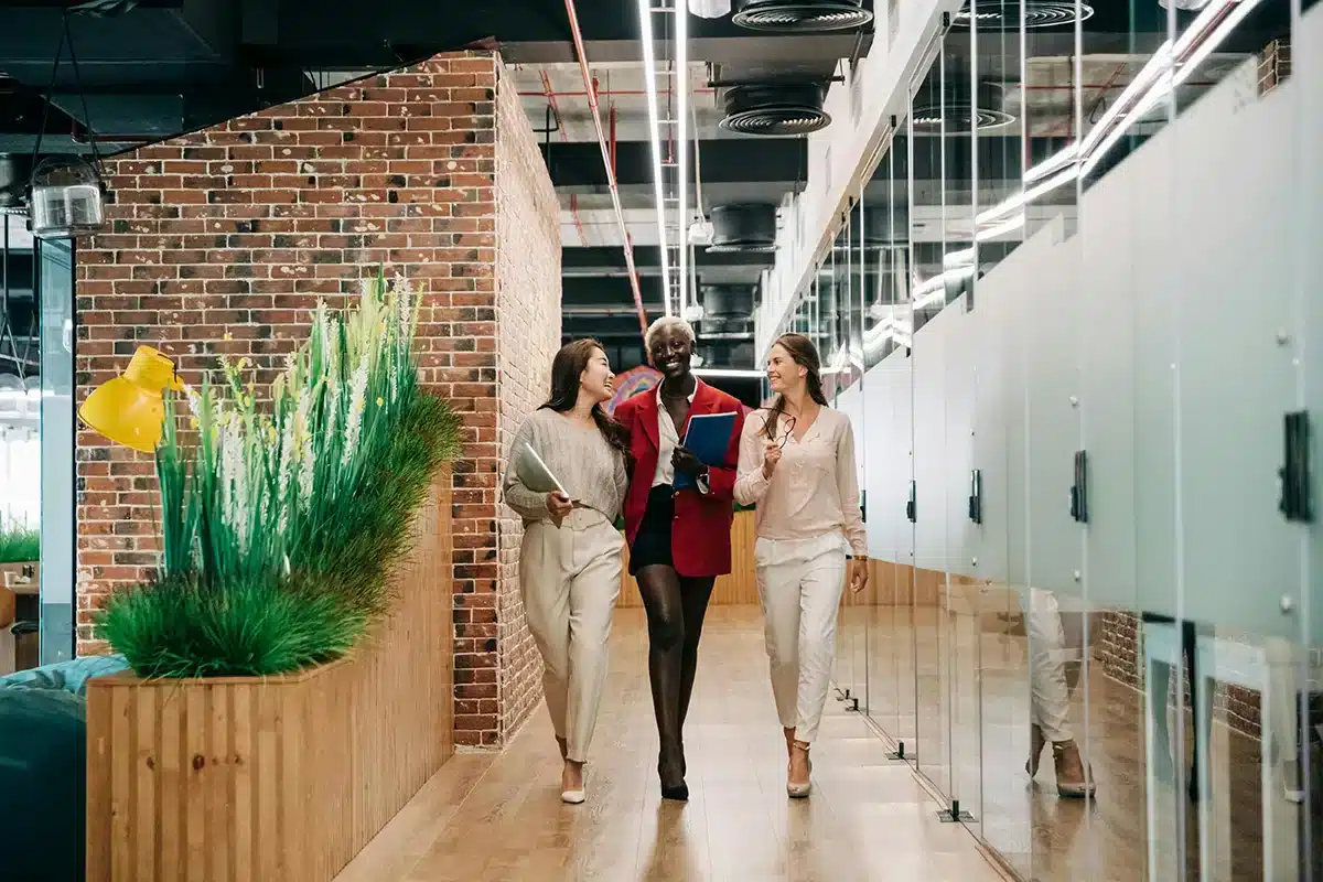 Three people walking down a modern office corridor with glass walls and brick accents. The person in the middle is wearing a red blazer and holding notebooks, while the other two are dressed in light-colored outfits. Theyre smiling and talking.