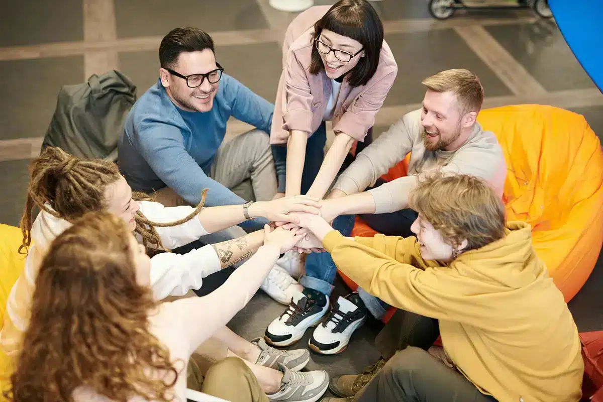 A group of seven people sit on colorful bean bags in a circle, smiling and stacking their hands together in the center. They appear to be in a brightly lit indoor space, engaging in a team-building exercise.