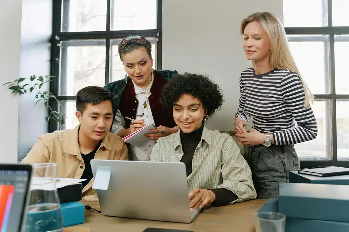 Four people are gathered around a table, looking at a laptop. One person is typing while the others watch and take notes. The room has large windows, and there are plants and office supplies on the table.