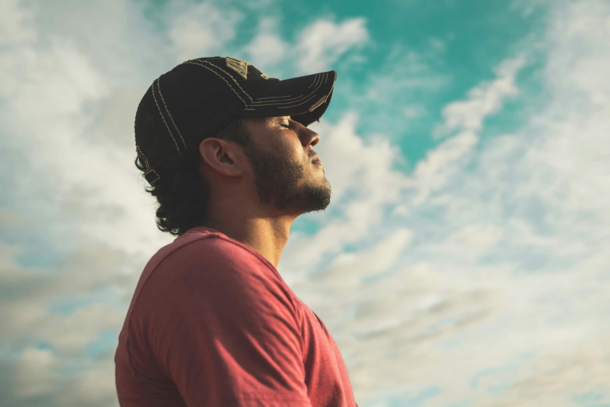 Man wearing a black cap with eyes closed under cloudy sky.