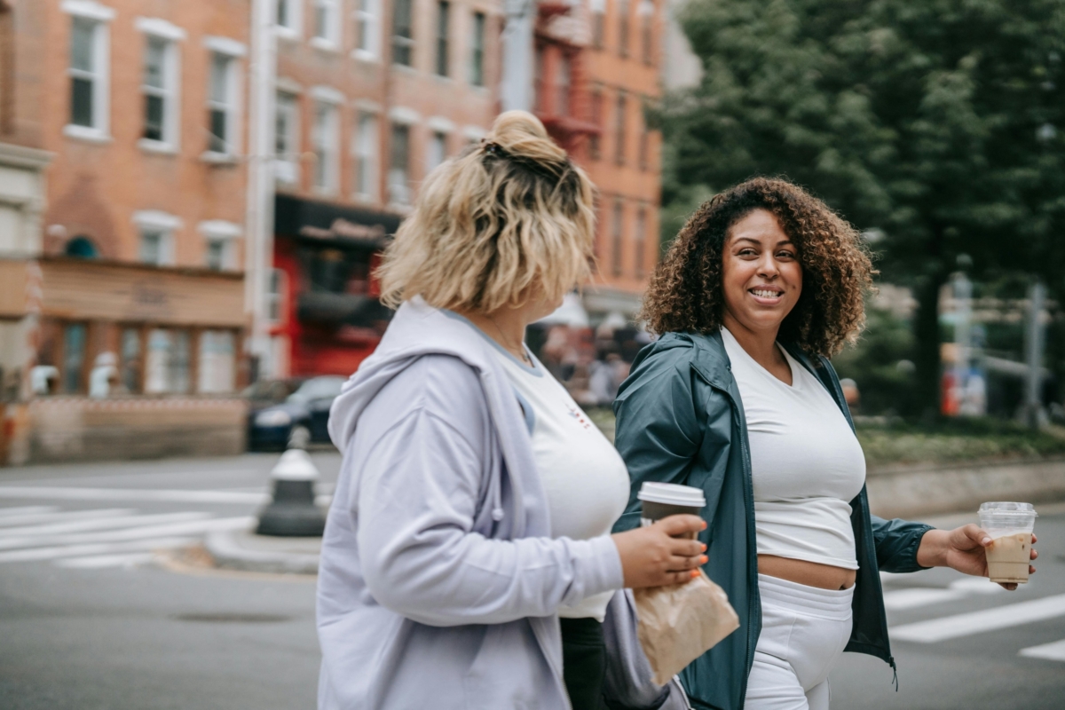 Two women smiling at each other and walking with a coffee and snack after a workout.
